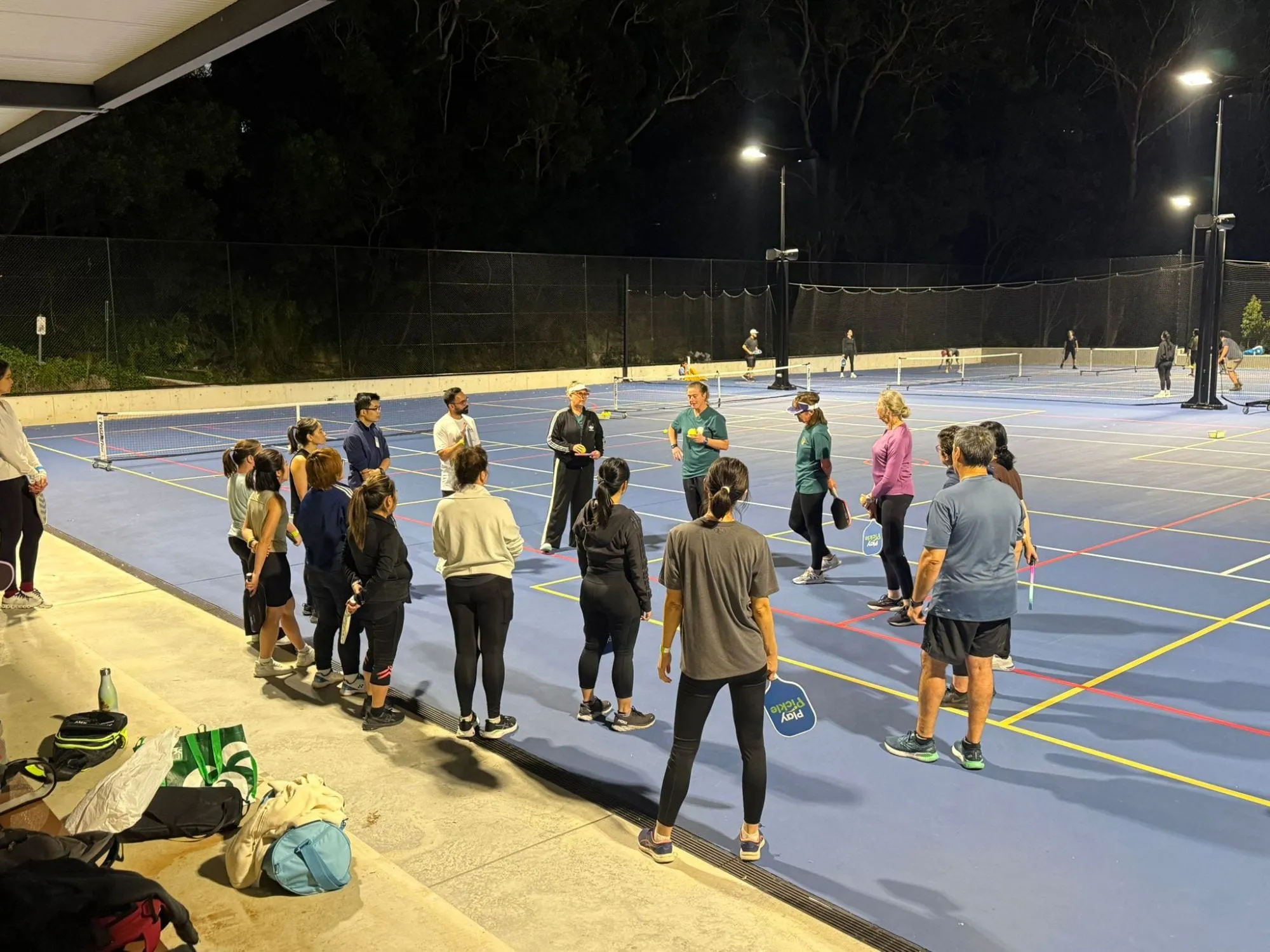 A large group of adult players gathered around two coaches on a floodlit court at an evening clinic