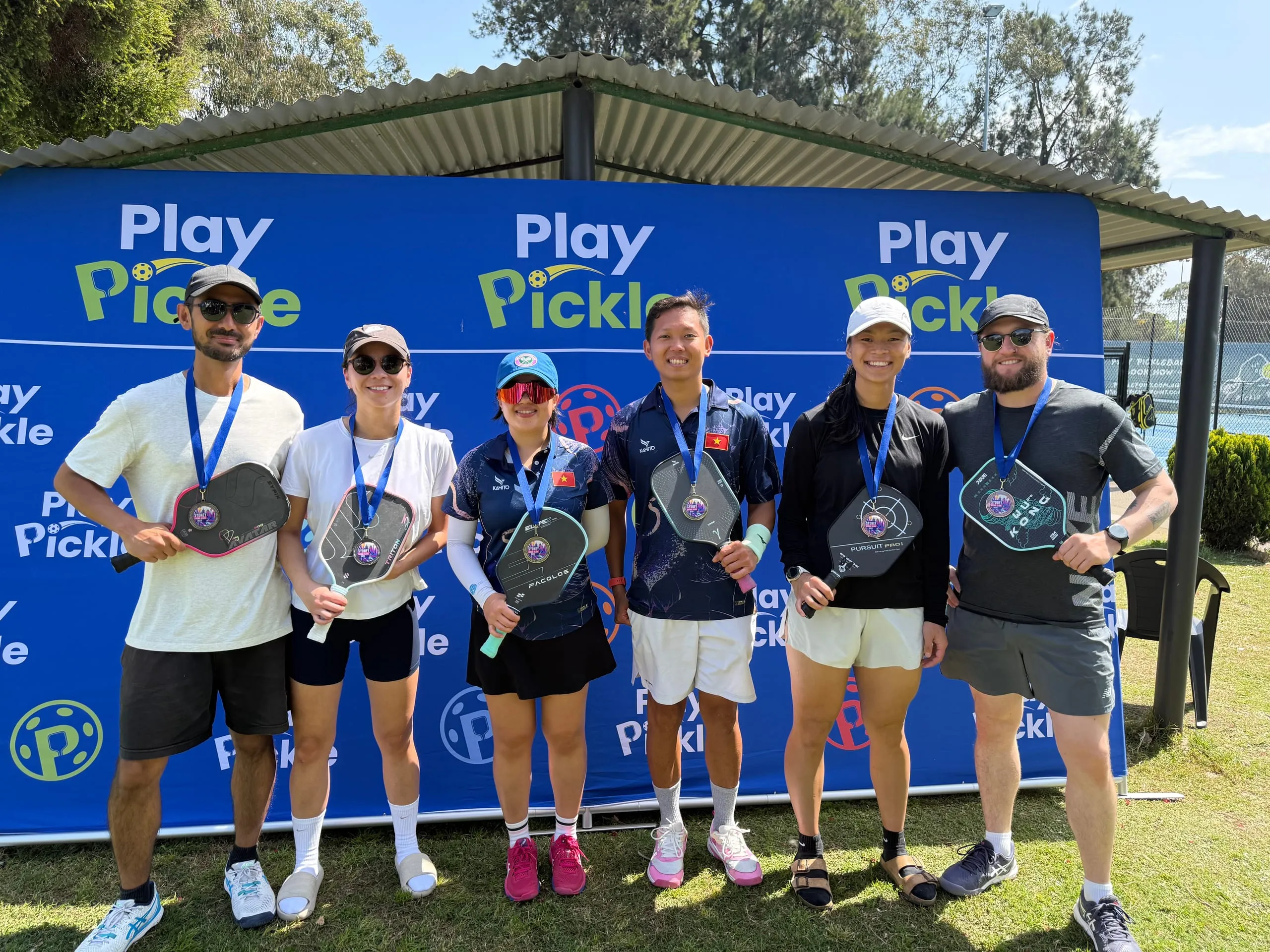 Six Play Pickle league players wearing medals and holding paddles in front of the Play Pickle banner