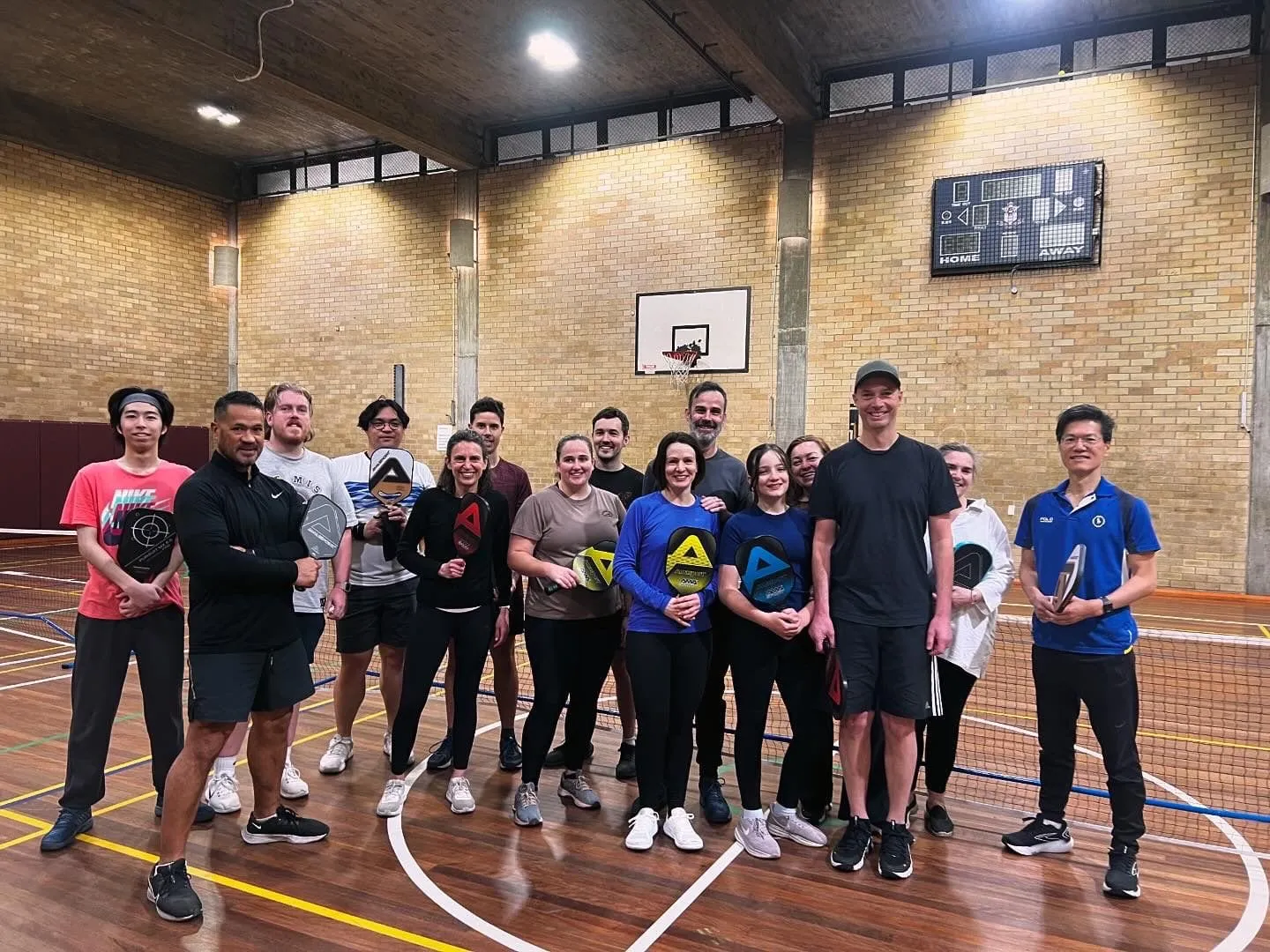 A social play group of about fifteen Play Pickle players standing together with their paddles on an indoor court